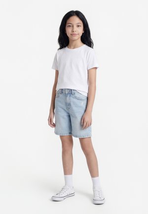 Ein Mädchen steht frontal zur Kamera und trägt ein weißes T-Shirt, hellblaue Jeansshorts, weiße Socken und weiße Turnschuhe vor einem schlichten weißen Hintergrund.