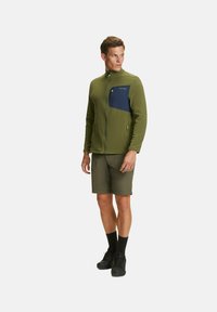 Giacca in pile verde con una tasca sul petto blu navy, maniche lunghe e colletto alto; indossata sopra pantaloni corti verdi e scarpe nere.