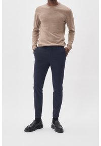 Beige lättviktskappa med rund hals, i kombination med marinblå slim-fit byxor och svarta loafers med en kraftig sula.