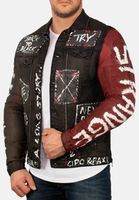 Veste en denim noir avec des manches à accents rouges, ornée de plusieurs patches peints et de textes en blanc. Texture usée et clous métalliques.