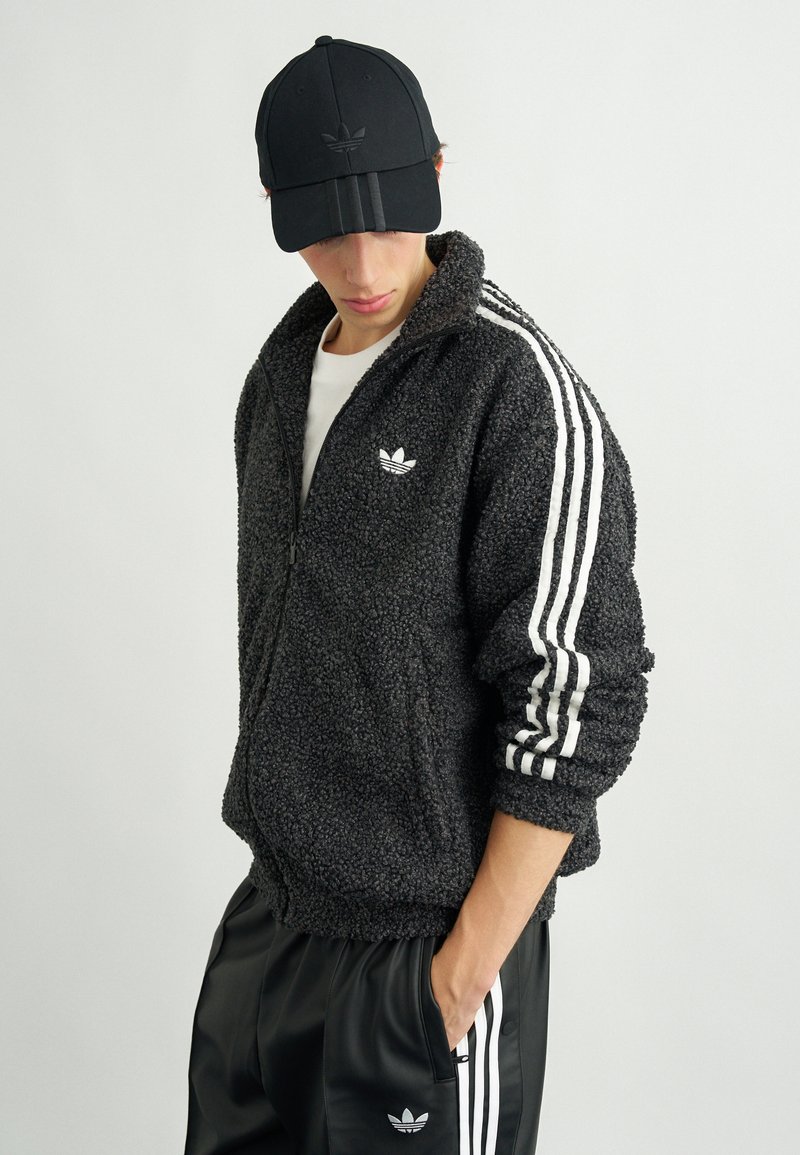 Μαύρο φουσκωτό μπουφάν με φερμουάρ και λευκές ρίγες στα μανίκια, με λευκό λογότυπο στο στήθος. Συνδυασμένο με μαύρα αθλητικά παντελόνια. Φοράει μαύρο καπέλο.