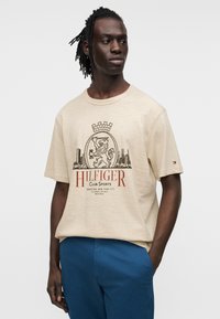 Μπέζ κοντομάνικο πουκάμισο με γραφικό σχέδιο σε σκούρο καφέ και κόκκινο, που απεικονίζει έναν λιοντάρι, κορώνα και αστικό ορίζοντα. Βαμβακερό ύφασμα, άνετη εφαρμογή.