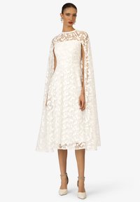 Witte kanten jurk met een hoge halslijn en vloeiende cape-mouwen. Bloemmotief, aansluitende lijfje en uitlopende rok. Eenvoudige beige hakken.