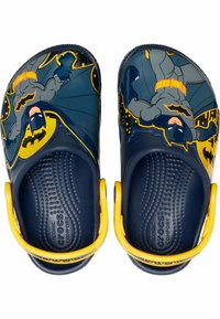 Des crocs bleus avec un design Batman et une sangle jaune. La surface est lisse avec des images détaillées de Batman contre un arrière-plan de la ville.