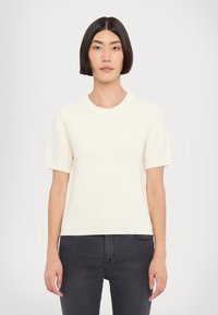 Kurzärmeliger cremefarbener Strickpullover mit gerippter Textur, runder Ausschnitt und lockerer Passform, kombiniert mit dunklen Jeans.