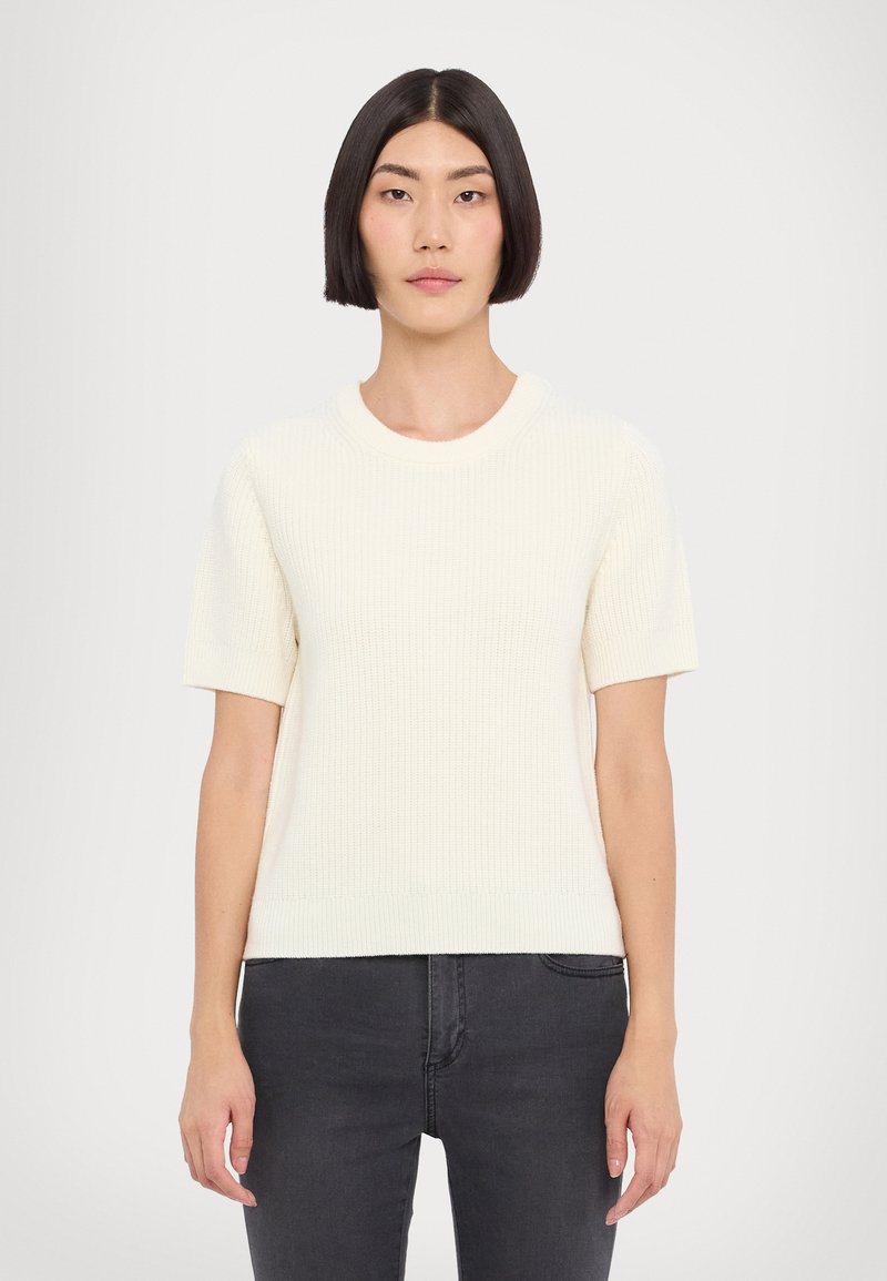 Kurzärmeliger cremefarbener Strickpullover mit gerippter Textur, runder Ausschnitt und lockerer Passform, kombiniert mit dunklen Jeans.