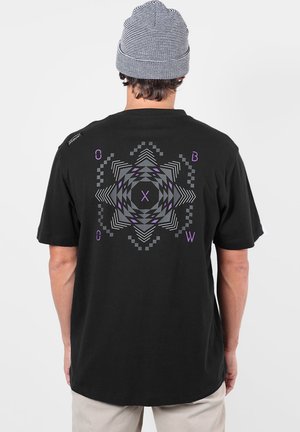Homme portant un t-shirt noir avec un motif étoile géométrique gris et violet et des lettres dans le dos, associé à un pantalon beige et un bonnet rayé.
