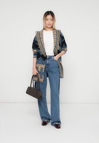 Cardigan împletit în nuanțe de albastru și bej, cu margini cu franjuri, modele tribale, asortat cu blugi deschiși la culoare și o geantă mică neagră.