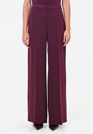 Pantaloni bordeaux a vita alta e gamba larga con dettaglio a bottone, indossati con scarpe bordeaux a punta e anello d'oro alla mano sinistra.