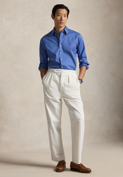 Homme portant une chemise bleue boutonnée, un pantalon blanc taille haute et des chaussures en cuir marron, debout les mains dans les poches.