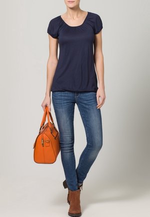 Femme debout portant un haut à manches courtes bleu marine, un jean bleu, des bottines marron et tenant un sac à main orange à côté d'elle.