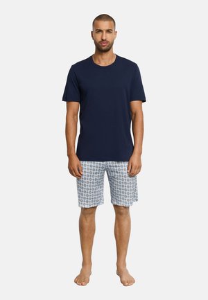 Hombre de pie descalzo, vistiendo una camiseta azul marino lisa y pantalones cortos hasta la rodilla de color azul claro con estampado, sobre un fondo blanco.