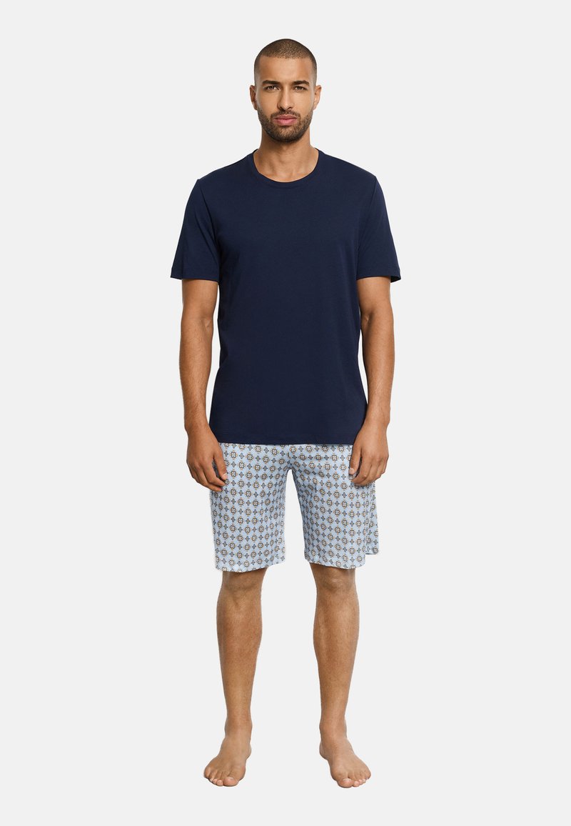 Mann steht barfuß und trägt ein einfaches marineblaues T-Shirt und hellblaue gemusterte knieumspielende Shorts vor weißem Hintergrund.