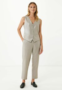 Gilet sans manches gris clair avec un décolleté en V et cinq boutons, associé à un pantalon droit assorti ; chaussures plates noires avec des clous argentés.