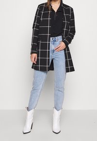 Femme portant un blazer à carreaux noirs, un haut zippé noir, un jean bleu clair et des bottes blanches à la cheville, se tenant devant un fond blanc.