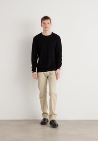 Un pull en maille noir, un pantalon beige clair et des chaussures noires. Le modèle se tient devant un fond blanc uni, affichant une tenue simple et décontractée.
