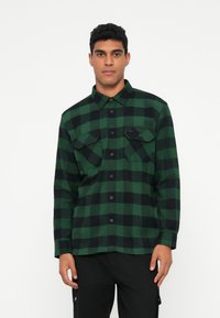 Camicia di flanella a quadri verdi e neri con maniche lunghe, chiusura con bottoni, due tasche sul petto e bottoni neri. Tessuto morbido.