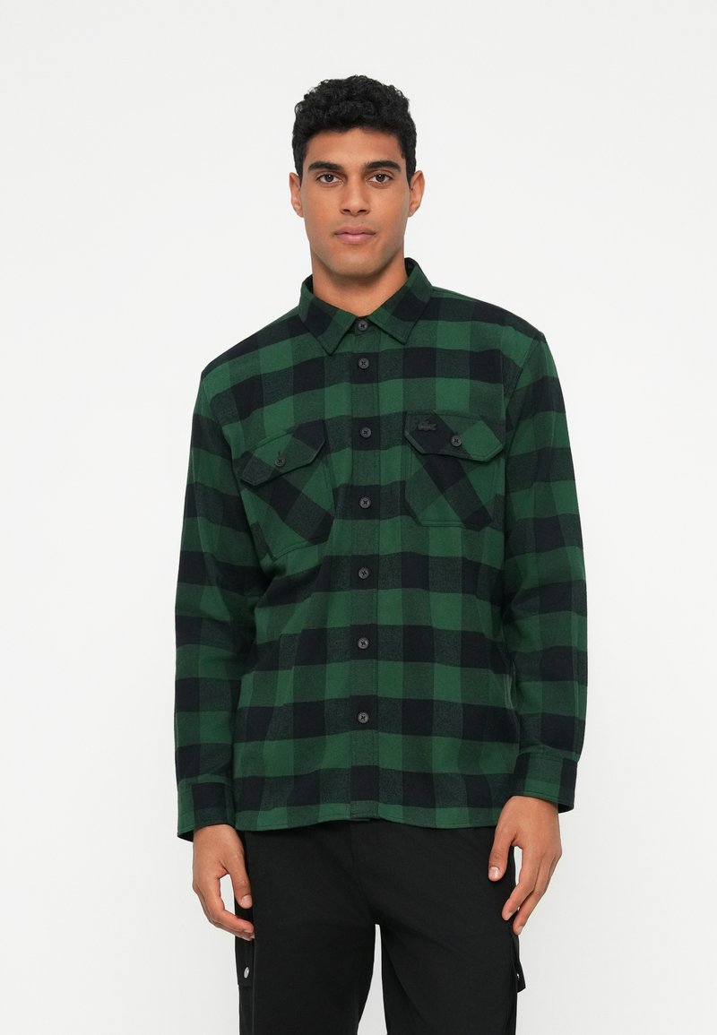 Camicia di flanella a quadri verdi e neri con maniche lunghe, chiusura con bottoni, due tasche sul petto e bottoni neri. Tessuto morbido.