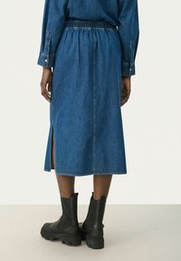 Zadní pohled na osobu v modré džínové midi sukni s postranním rozparkem a černými robustními kotníkovými botami na světlém pozadí.