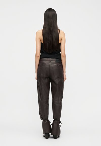 Γυναίκα με μακριά σκούρα μαλλιά στέκεται με την πλάτη προς τα εμάς, φοράει μαύρο αμάνικο τοπ και ψηλόμεσα σκούρα καφέ δερμάτινα παντελόνια με κορδόνια στον αστράγαλο.