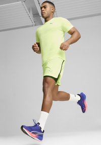 Camiseta y pantalones cortos atléticos de color amarillo neón con acentos en verde oscuro, combinados con zapatillas de correr azules y rosas, llevadas con calcetines blancos.
