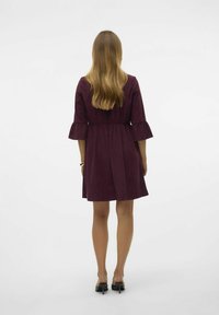 Maroon obleka s širokimi rokavi, izdelana iz mehke tkanine. Ima združeno pasnico in do kolen dolžino, kombinirana z črnimi petkami.