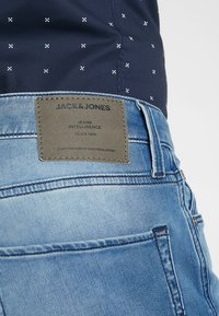 Jasnoniebieskie dżinsy z widocznym szwem, skórzana metka z napisem "JACK & JONES" oraz ciemna koszula w wzory częściowo widoczna powyżej.