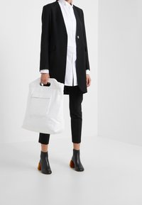 Vit tote-väska med handtag, strukturerad yta, tillsammans med en svart kavaj, vit skjorta med veck och svarta ankelskorsetter med bruna klackar.