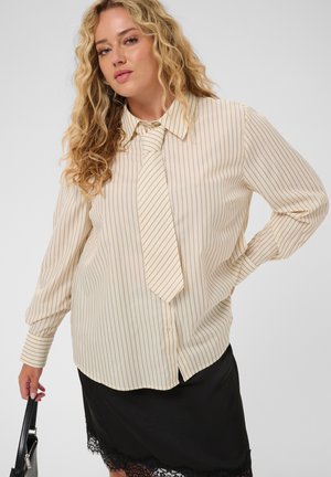Femme aux cheveux blonds bouclés portant une chemise beige rayée et une cravate, jupe noire avec ourlet en dentelle, tenant un sac à main noir d'une main.