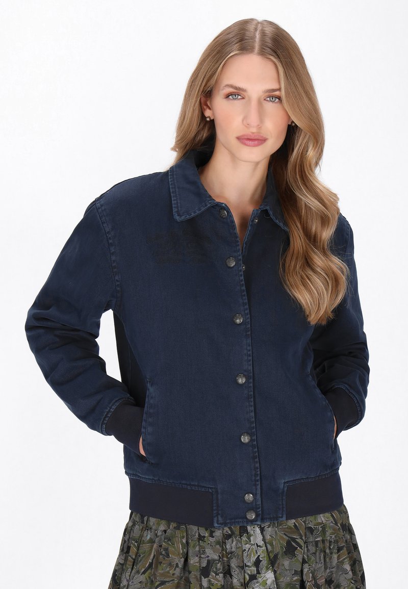 Tengerészkék farmerdzseki gombos elejével, bordás mandzsettákkal és zsebekkel; gallérral és sima textúrával rendelkezik.