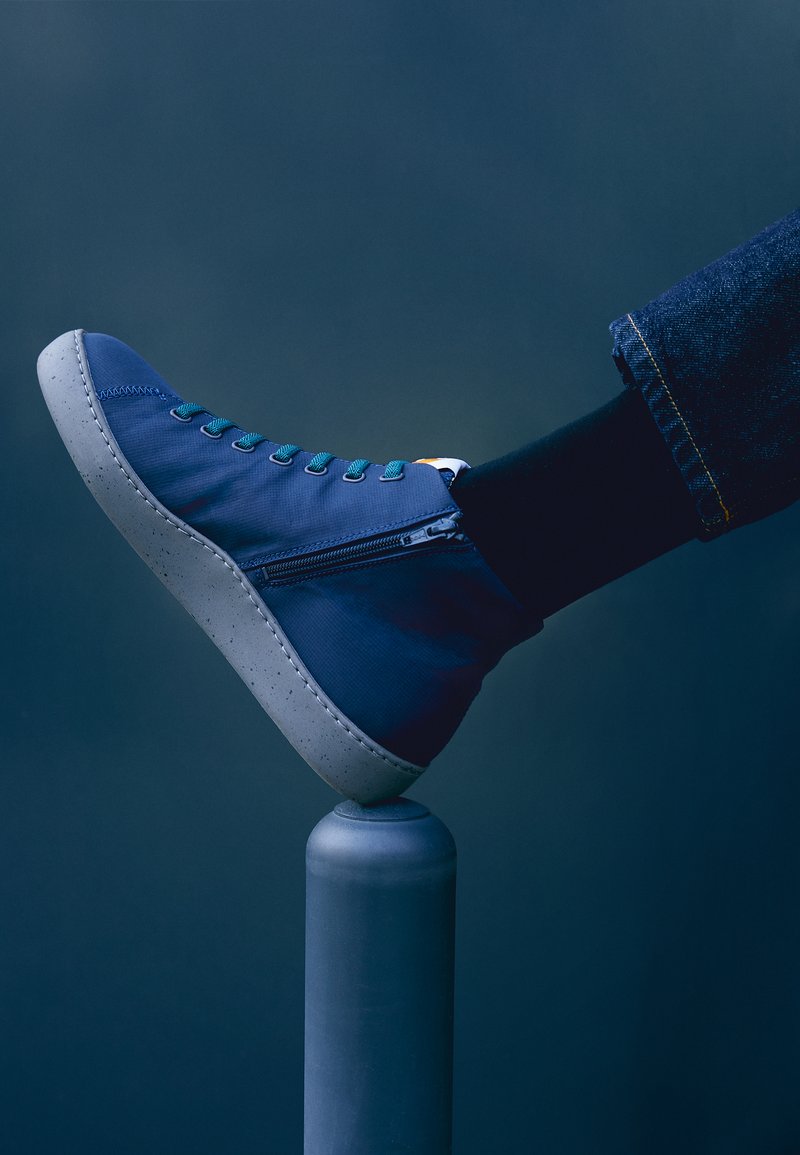 Høje, marineblå sko med en tekstureret overdel, teal snørebånd, en sidelukning med lynlås og en gråspraglet sål. Placeret på en cylindrisk støtte.