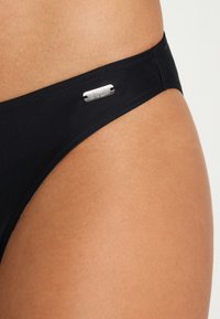 Schwarzer Bikini-Unterteil mit minimalistischem Design, ausgestattet mit einem silbernen Etikett mit der Aufschrift "Venice Beach" an der Seite, glatte Textur.