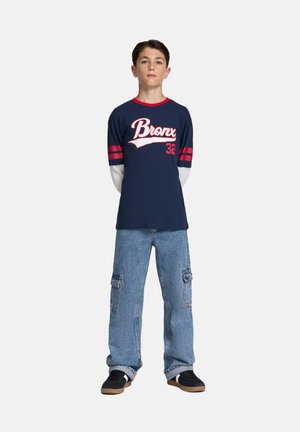 T-shirt blu navy con stampa bianca "Bronx", dettagli rossi e maniche lunghe grigie. Abbinata a jeans cargo azzurri chiari e sneakers nere.