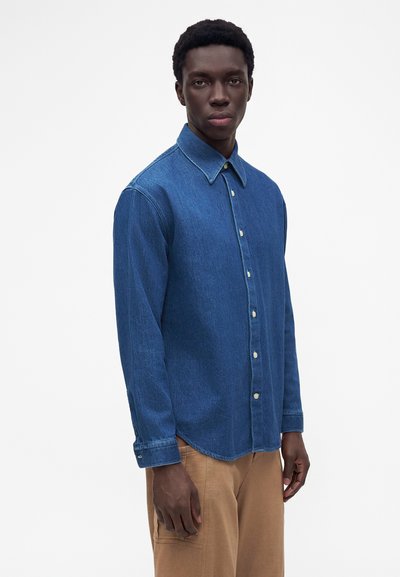 Jeune homme portant une chemise en denim bleue à manches longues boutonnée et un pantalon marron clair, debout devant un fond blanc uni.