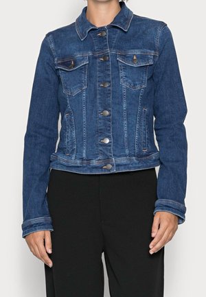 Frau trägt eine taillierte blaue Jeansjacke mit Knopfleiste und Klappentaschen auf der Brust, kombiniert mit schwarzen Hosen.