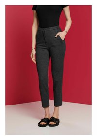 Žena v tmavě šedých kalhotách vysokého pasu, černém krátkém topu, černých sandálech, se zlatým náramkem a prstenem, stojí na pozadí červené a bílé.