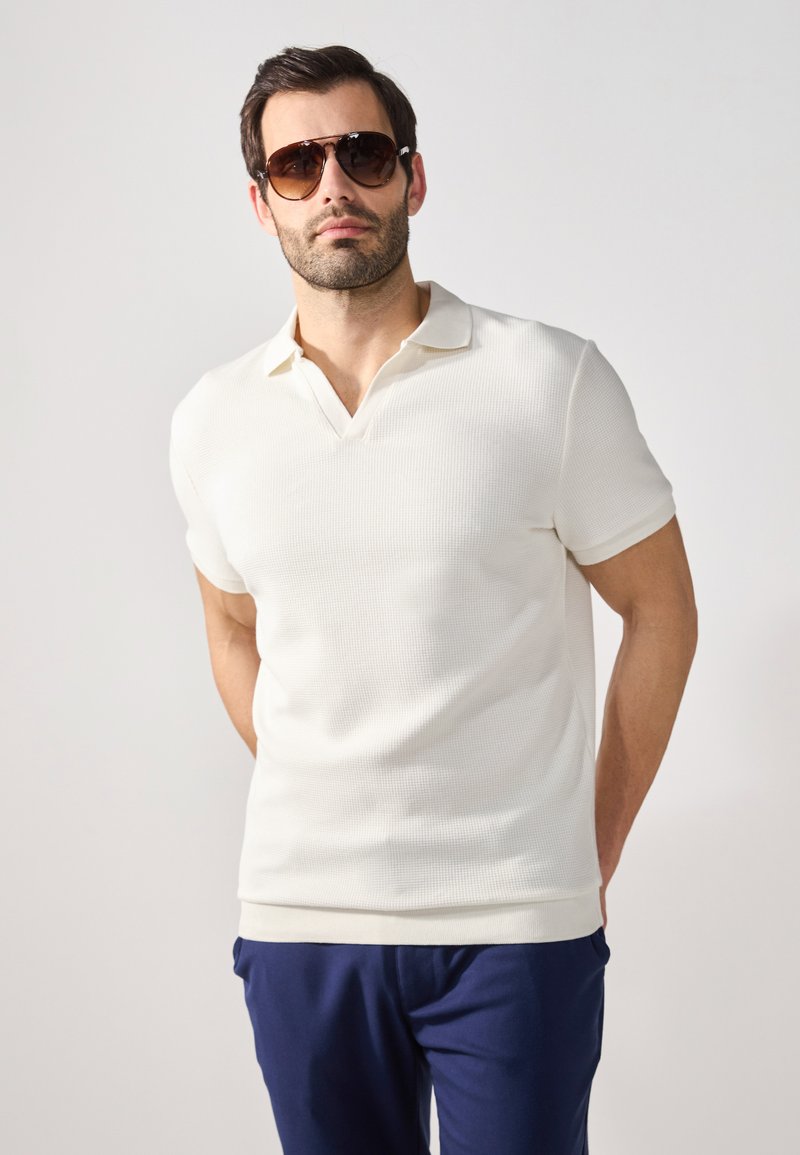 Weißes Poloshirt mit kurzen Ärmeln aus strukturiertem Stoff und V-Ausschnitt, kombiniert mit marineblauen Hosen. Das Modell trägt eine Sonnenbrille.