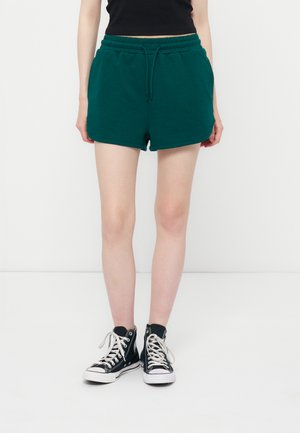 Kobieta ubrana w ciemnozielone szorty ze ściągaczem, czarne wysokie trampki z białymi sznurówkami i czarny krótki top, stojąca na białej podłodze.