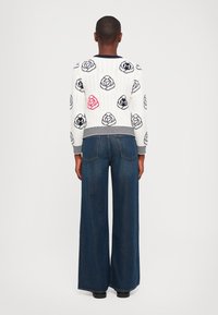 Strikkegenser i hvit med kabelstruktur, mønstrete med svarte og røde roser, kombinert med vide mørk denimbukser.