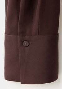Poignet en tissu marron foncé avec un seul bouton assorti et des détails de couture visibles sur un vêtement à manches longues.