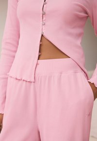 Zamatá, svetloružová súprava loungewear obsahujúca top s dlhým rukávom a zapínaním na gombíky a bočným výrezom, doplnená elastickými nohavicami s logom.