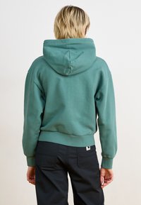 Grønn hettegenser med cropet passform, ribbede mansjetter og kant. Myk, glatt tekstur med et enkelt design, brukt med mørke bukser.