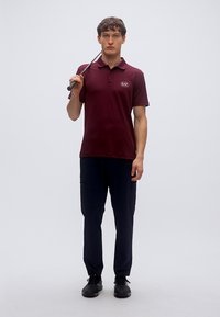 Μπορντό πόλο πουκάμισο με γιακά και λογότυπο, συνδυασμένο με σκούρο παντελόνι cargo. Το μοντέλο κρατά ένα γκολφ κλαμπ, φοράει μαύρα παπούτσια και βρίσκεται σε ουδέτερο φόντο.