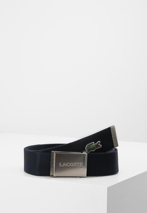 Navy blue fabric belt with silver buckle displaying "Lacoste" logo and a small embroidered green crocodile near the end of the belt.