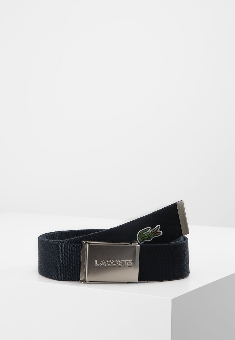 Ceinture en tissu bleu marine avec boucle argentée affichant le logo "Lacoste" et un petit crocodile vert brodé près de l'extrémité de la ceinture.