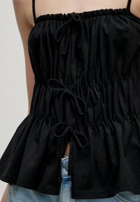 Zwarte katoenen top met geplooid textuur, verstelbare trekkoorden en een uitlopende zoom. Gecombineerd met blauwe denim shorts met een metalen knoop.