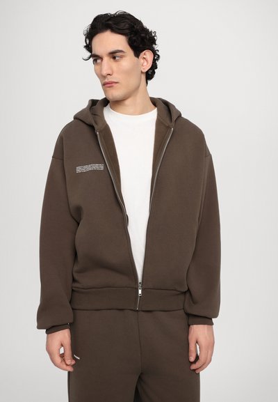 Rjav zip-up kapucar iz materiala, ki se uporablja za izdelavo puloverjev. Ima sprednji žep in belo besedilo na levi strani. Gladka tekstura.