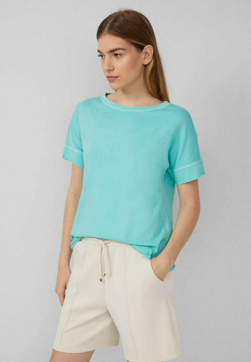 Lichtblauwe t-shirt met korte mouwen, gemaakt van zacht materiaal, met een losse pasvorm en contrasterende stiksels. Gecombineerd met beige shorts.