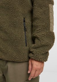 Olive rohelise fleece-jakk, millel on tekstuurne, hõrk pind, varustatud lukuga küljetasku ja kontrastset kangast varrukad.