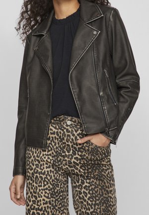 Schwarze Kunstleder-Bikerjacke mit silbernen Reißverschlüssen und Druckknöpfen, kombiniert mit beigen Hose im Leopardenmuster. Die Jacke hat einen Reverskragen und Reißverschlusstaschen.